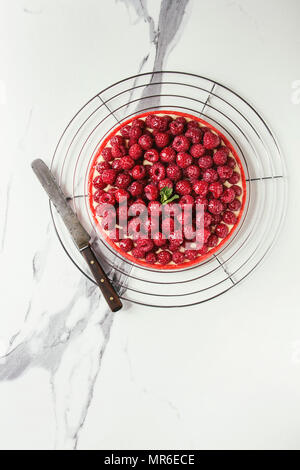 Tarte sablée framboise rouge et crème de citron framboises fraîches servi glacé sur grille de refroidissement avec couteau sur du marbre blanc texture background. Banque D'Images