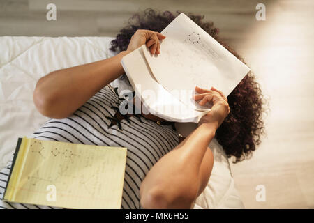 African American college student faire leurs devoirs au lit à la maison. Femme noire en chimie des formules, des problèmes à mémoriser des notes sur l'exercice Banque D'Images