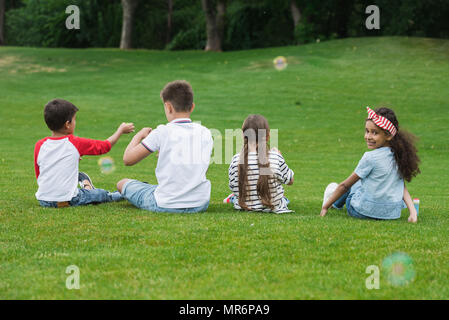 Vue arrière d'adorables enfants jouant avec multiethnique des bulles de savon en étant assis sur l'herbe verte dans le parc Banque D'Images