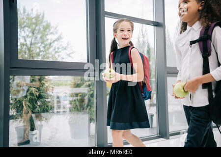 Smiling holding apples écolières multiethnique tout en marchant dans le couloir de l'école Banque D'Images