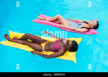 Les jeunes femmes multiethniques flottant sur le matelas gonflable dans la piscine du resort Banque D'Images