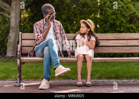 Petite-fille afro-américaine avec bras croisés à la recherche de son grand-père en conversation sur smartphone dans park Banque D'Images