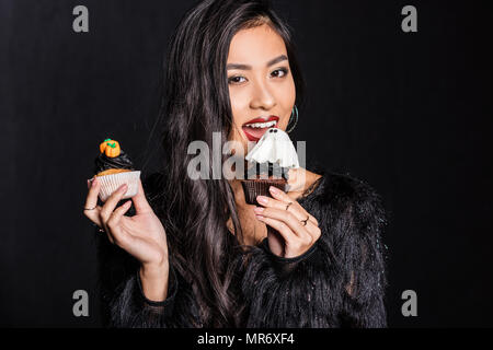 Belle asiatique femme en manteau de plumes d'autruche tenant deux cupcakes dans ses mains et looking at camera Banque D'Images