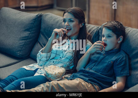 Petit-fils et sa jeune mère assis sur la table pendant que vous regardez la télévision et eating popcorn Banque D'Images