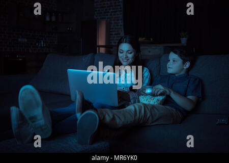 Petit garçon et sa belle mère regarder la vidéo sur ordinateur portable et eating popcorn, alors qu'il était assis sur la table Banque D'Images