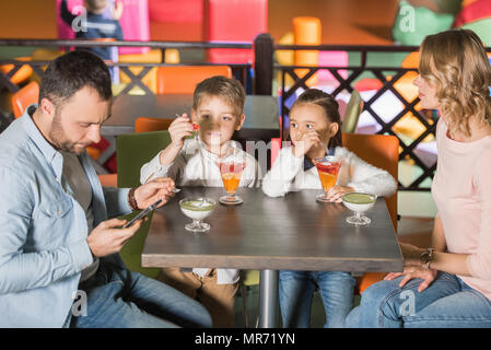 Famille avec deux enfants de manger savoureux desserts et le père à l'aide de centre de divertissement de smartphone Banque D'Images