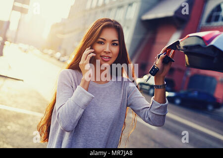 Young Asian business woman with phone debout à la voiture avec les clés sur l'arrière-plan de la ville en matinée Banque D'Images