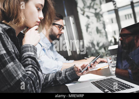 Young businesswoman avec des lunettes et un téléphone intelligent à la main fonctionne avec ordinateur portable. Travailler avec les appareils numériques dans l'espace loft moderne Banque D'Images