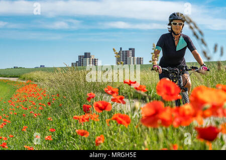 Nice senior woman riding a e-Mountainbike dans la banlieue d'une grande ville, entourée de champs verts et rouge coquelicot Banque D'Images