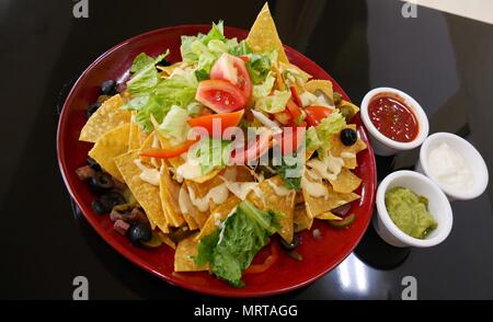 Nachos garnis de tomates, laitue, olives et lardons dans une plaque ronde rouge, servi avec des petits bols de ketchup, mayonnaise et avocat rafraîchir Banque D'Images