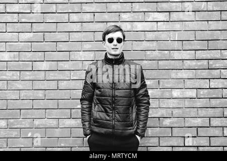 Un jeune homme est appuyé contre un mur de brique en lunettes image en noir et blanc. Banque D'Images