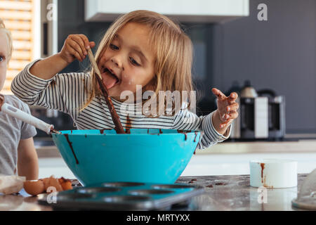 Petite fille léchant tout en mélangeant à la cuillère la pâte pour la cuisson dans la cuisine et son frère par permanent. Cute little children making pâte pour la cuisson. Banque D'Images