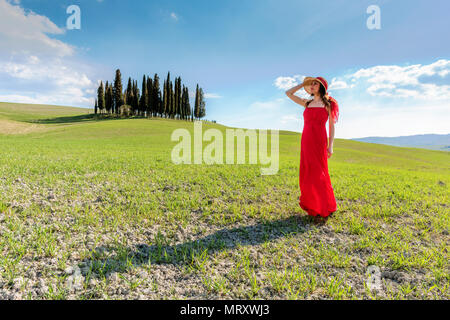 San Quirico d'Orcia, Val d'Orcia, Sienne, Toscane, Italie. Une jeune femme en robe rouge à admirer la vue dans un champ de blé près du cyprès de l'Orcia va Banque D'Images