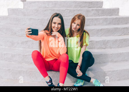 Deux amies dans des tenues de sport ne une assise sur les marches selfies à l'extérieur. Les deux jeunes femmes heureux et souriant à la détente dans un Banque D'Images