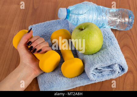 La main féminine avec haltère jaune et vert frais et une bouteille d'eau d'apple sur bleu serviette sur plancher en bois. L'alimentation, le sport, la condition physique et en bonne santé concept Banque D'Images