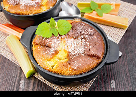 Clafouti tarte à la rhubarbe dans un pot de noir, de menthe et de tiges de rhubarbe sur une serviette en osier sur fond de planche de bois foncé Banque D'Images