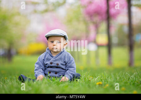 Portrait of cute smiling élégant tout-petit garçon assis n l'herbe dans le parc à l'extérieur. Beau garçon souriant positives blonde wearing blue shirt et télévision ca Banque D'Images
