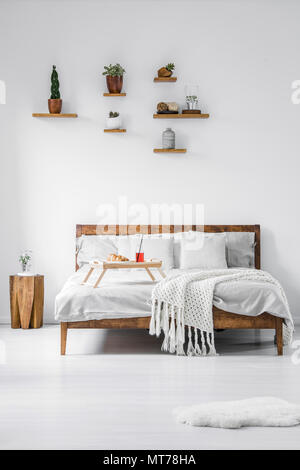 Vue de face d'un lit double en bois avec des draps, oreillers et couvertures dans une chambre contemporaine de l'intérieur de l'hôtel. Bac alimentaire stand avec petit déjeuner Banque D'Images