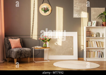 Petite table avec livre ouvert et tulipes fraîches debout à côté de fauteuil gris avec coussin dans un décor intérieur avec salle de lecture et de peintures Banque D'Images