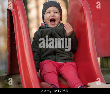 Happy Baby Boy avec un rire très fort les enfants assis sur une diapositive à l'aire de jeux pour enfants. Concept d'enfance active Banque D'Images