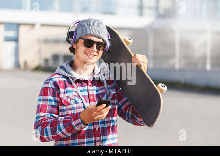 Hipster homme avec lunettes habillé en chemise et le chapeau cheked holding smartphone et du skateboard, écoute la musique concept Banque D'Images
