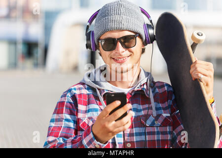 Hipster homme avec lunettes habillé en chemise et le chapeau cheked holding smartphone et skateboard à écouter de la musique Banque D'Images