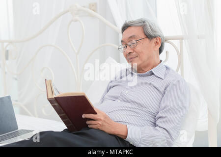 Vieil homme asiatique professeur lunettes lecture du texte livre chaque matin sur le lit Banque D'Images