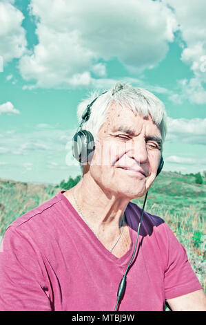 Un homme âgé aux yeux clos dans les écouteurs écoute de la musique tonique photo. Personnes âgées smiling man sitting extérieur sous ciel nuageux. L'influence de la musique sur le cerveau Banque D'Images