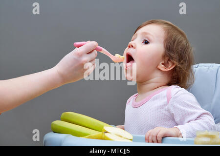 Un peu jolie fille est alimenté par sa mère avec une cuillère pleine de purée de fruit, assis sur la chaise d'alimentation gris et blanc sur le fond gris. Studio shot Banque D'Images