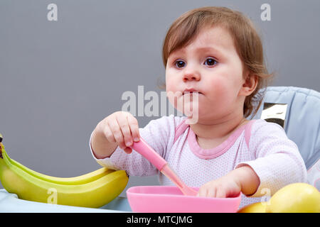 Un peu jolie fille est alimenté par sa mère avec une cuillère pleine de purée de fruit, assis sur la chaise d'alimentation gris et blanc sur le fond gris. Studio shot Banque D'Images