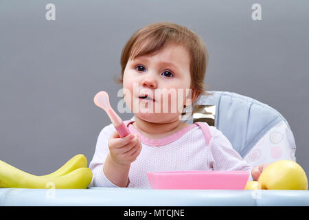 Un peu jolie fille est alimenté par sa mère avec une cuillère pleine de purée de fruit, assis sur la chaise d'alimentation gris et blanc sur le fond gris. Studio shot Banque D'Images