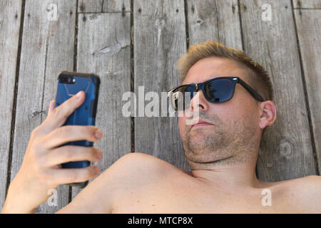 Nude young 40 caucasian man using mobile phone smartphone sur patio où se détendre dans le soleil Banque D'Images