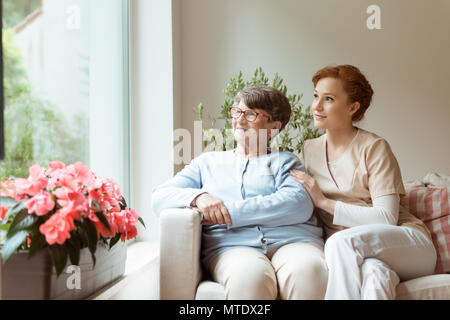 Geriatric femme et son gardien professionnel assis sur un canapé et regarder à travers une fenêtre dans une salle de séjour. Fleurs sur un rebord de fenêtre. Banque D'Images