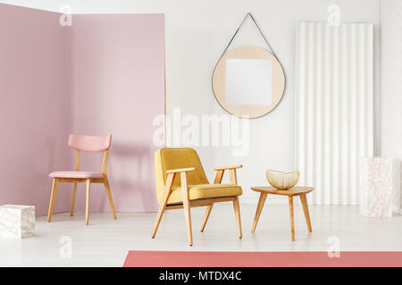 Chaise rose dans l'angle et fauteuil jaune placés par une table en bois avec bol d'or dans cette chambre lumineuse intérieur avec miroir sur le mur Banque D'Images