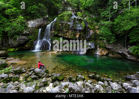 Woman in red jacket assis sur un gros rocher en face de Karlovac waterfal, vallée de la soca, Bovec, Slovénie Banque D'Images
