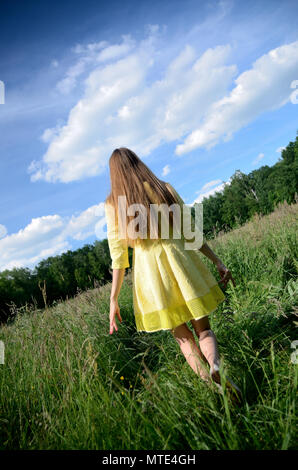 Photo de derrière sans visage. Jeune femme robe jaune avec balades bénéficie d'sur l'herbe. Verte prairie de fleurs sauvages et de ciel bleu. Banque D'Images