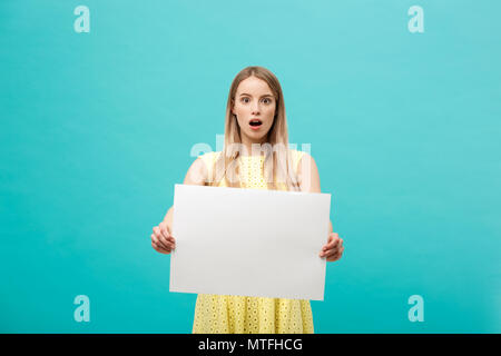 Portrait de jeunes surpris blond woman holding blank sign with copy space on blue background studio. Montrant choqué face surprise. Banque D'Images