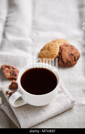 Le café du matin dans la tasse de porcelaine blanche, croustillant de chocolat cookies sur homespun serviette sur nappe blanche, close-up, selective focus, vue de côté. Banque D'Images