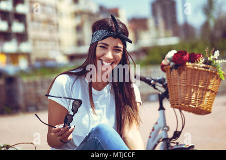 Happy smiling young woman is enjoying in a été ensoleillé jour, assis sur un banc de la ville, à côté du vélo avec panier de fleurs. Banque D'Images