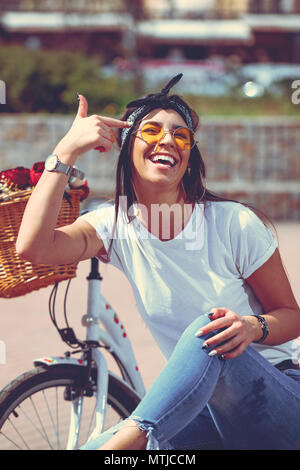 Jolie jeune femme est heureux de s'amuser et profiter de mettre un doigt sur son front, dans une journée ensoleillée d'été, à côté du vélo avec panier de fleurs. Banque D'Images