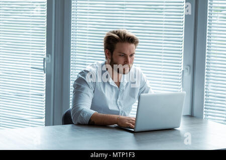 Jeune homme sérieux travaillant sur un ordinateur portable avec un look de concentration assis à une table en face de grandes fenêtres avec stores Banque D'Images