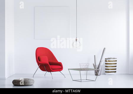 Repose-pied en osier rond placé sur le sol à côté d'un fauteuil rouge blanc en salle d'attente avec table et abat-jour industriel Banque D'Images