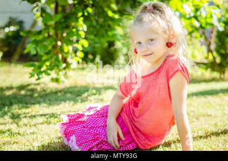 Adorable petite fille blonde avec la cerise dans les oreilles assis dans l'herbe verte Banque D'Images