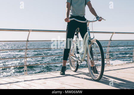 Jeune femme élancée reposant sur un vintage bicycle sur le bord de l'eau. Femme debout avec un sac à dos lors d'une promenade en vélo sur le fond de la mer le soleil Banque D'Images