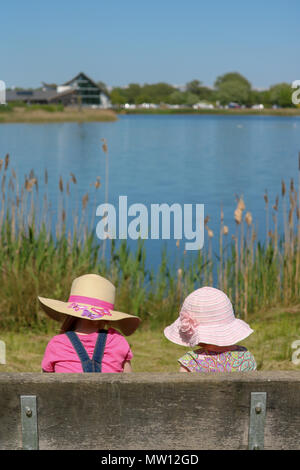 Une jeune fille et un bébé, dos à la caméra et à la fois dans sunhats, s'asseoir sur un banc et contempler la vue sur un lac sur un jour ensoleillé à Stanwick Lacs, Northampton, Royaume-Uni Banque D'Images