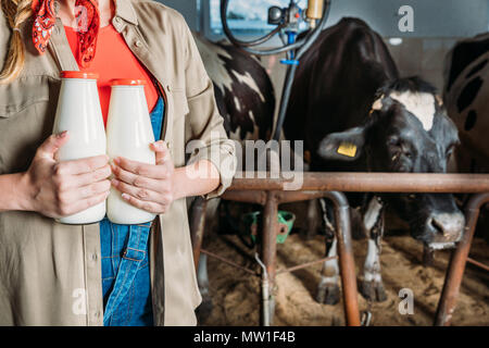 Cropped shot of female farmer holding bouteilles avec du lait frais en étant debout près de vaches dans stall Banque D'Images