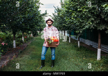 Agriculteur barbu en chemise à carreaux et hat holding venu des légumes en panier et looking at camera Banque D'Images