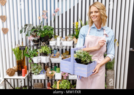 Smiling young female florist holding avec panier en bois et des plantes à l'écart dans le magasin de fleurs Banque D'Images