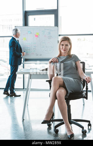 Belle businesswoman sitting on chair and smiling at camera while senior businessman standing near tableau blanc derrière Banque D'Images