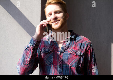 Portrait of attractive guy faisant appel téléphonique avec arrière-plan uni. Avec l'homme portant la barbe flanelle parlant au téléphone au soleil venant de la fenêtre. Banque D'Images